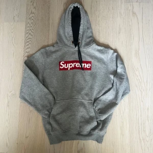 Grå Supreme hoodie med box logo - Grå hoodie från Supreme med ikonisk röd box logo på bröstet. Tröjan har huva med svart snörning och stor magficka framtill. Mjuk insida och ribbade muddar vid ärmslut och nederkant. Perfekt för dig som gillar streetwear och vill sticka ut.