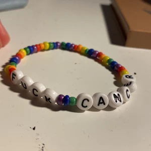 Två färgglada armband med pärlor och snäcka - Säljer två unika armband! Ett med regnbågsfärgade pärlor och vita bokstavspärlor med texten 'FUCK CANCER'. Det andra har rosa snöre, silverfärgade pärlor, ljusrosa stenar och en snäcka med rosa prickar. Perfekta accessoarer för dig som vill sticka ut.25kr styck eller 30 för båda!