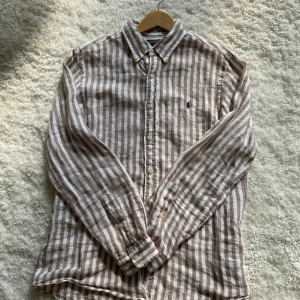 Randig linneskjorta från Polo Ralph Lauren - Snygg randig skjorta från Polo Ralph Lauren i brun och vit. Skjortan är i linne, har klassisk krage, långa ärmar och knappar framtill. Custom fit-modell med broderad logga på bröstet. 