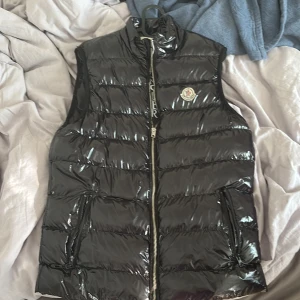 Svart dunväst från Moncler - Säljer en svart, glansig dunväst från Moncler med hög krage och dragkedja framtill. Västen har två fickor med dragkedja och klassisk Moncler-logga på bröstet. Perfekt för lager-på-lager och riktigt snygg till streetwear eller casual outfits.