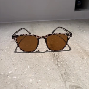 Leopardmönstrade solglasögon brun - Snygga solglasögon med leopardmönstrad båge i plast och bruna glas. Runda linser och klassisk form som ger en cool vibe. Perfekta för att lyfta din stil under soliga dagar.