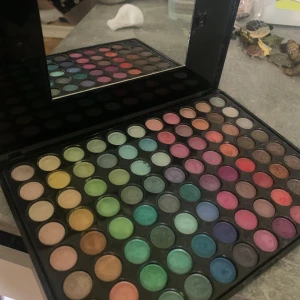 Stor ögonskuggspalett med spegel - En stor ögonskuggspalett med 88 färger i både matta och skimrande nyanser. Paletten innehåller allt från neutrala bruna och beige toner till färgstarka blå, gröna, rosa, lila och gula nyanser. Kommer i svart plastfodral med inbyggd spegel. Tydliga tecken på användning men många av färgerna har inte rörts. Locket har tyvärr ramlat av (går inte att stänga ordentligt). 