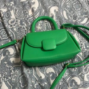 Grön handväska från Primark - Snygg grön handväska från Primark med stilren design och guldfärgade detaljer. Väskan har ett stadigt handtag, avtagbar axelrem och praktiskt innerfack. Tillverkad i syntetmaterial med slät finish och enkel knäppning framtill.