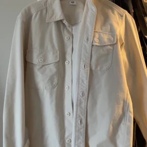 Off white overshirt från Uniqlo - Stilren off white overshirt från Uniqlo med klassisk krage, två bröstfickor med lock och knappar framtill. Skjortan är långärmad och tillverkad i mjuk bomull, perfekt att bära öppen eller stängd för en avslappnad look. Xs men sitter mer som en S