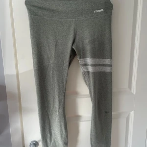 Grå leggings från Stronger - Snygga grå leggings från Stronger med vita ränder på ena benet. Hög midja och tight passform, perfekta för träning eller chill. Mjukt och stretchigt material i polyester och elastan som sitter skönt hela dagen.