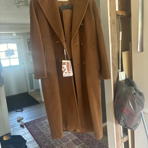 Max Mara brun kappa i ull, stl 36 - Snygg lång kappa från Max Mara i en varm brun tobaksnyans. Klassisk dubbelknäppt design med breda slag, fickor på sidorna och tillhörande bälte. Tillverkad i mjuk ull som känns lyxig och tidlös. Perfekt för dig som vill ha en stilren och elegant look.
