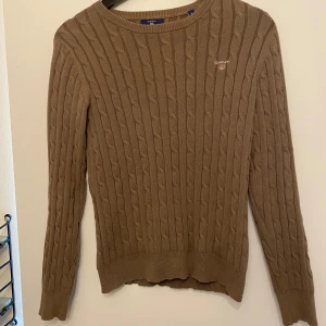 Beige kabelstickad tröja från GANT - Säljer en beige kabelstickad tröja från GANT i stretchig bomull. Tröjan har rund halsringning, lång ärm och diskret GANT-logga broderad på bröstet. Perfekt för lager-på-lager och snygg till både jeans och kjol.
