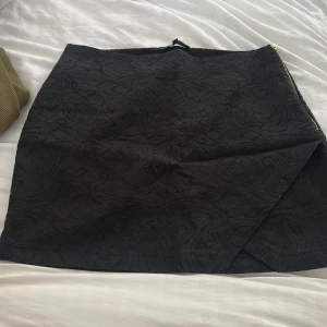 Svart omlottkjol med strukturmönster - Snygg svart kortkjol med omlottdesign och subtilt strukturmönster. Kjolen har en guldfärgad dragkedja i sidan som detalj. .