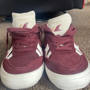 Bordeaux sneakers från Pepe Jeans - Snygga sneakers från Pepe Jeans i bordeauxröd mocka med vita och grå detaljer. Skorna har vit sula, snörning och logga på plösen. Baksidan har grå mocka och siffran 1973. Perfekta för dig som gillar en sportig och stilren look.