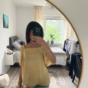 Ljusgul blus med fyrkantig ringning - Säljer en ljusgul blus med korta, luftiga ärmar och snygg fyrkantig ringning. Blusen har en lös passform och är tillverkad i ett glansigt, silkesliknande material som känns lyxigt och svalt. Perfekt för dig som gillar en stilren och fräsch look.