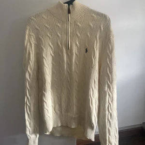 Vit kabelstickad tröja Polo Ralph Lauren - Kabelstickad vit tröja från Polo Ralph Lauren med half zip och hög krage. Klassisk design med broderad logga på bröstet. Tillverkad i mjuk bomull och har långa ärmar. Perfekt för dig som gillar preppy och stilren look.