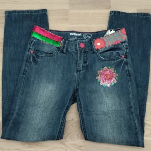 Desigual jeans med paljetter - Jeans från Desigual med färgglada detaljer som paljettbälte i rosa och grönt, broderad blomma med paljetter på benet och mönstrad linning. 