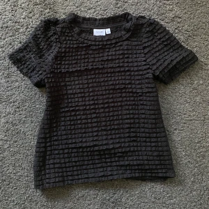 Svart ribbad topp från Vila XS - Svart topp från Vila i storlek XS med korta ärmar och ett unikt ribbat strukturmönster över hela plagget. Toppen har rund halsringning och är tillverkad i ett lätt och luftigt material.