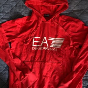 Röd EA7 hoodie från Emporio Armani - Snygg röd hoodie från EA7 Emporio Armani med vit logga framtill. Tröjan har huva med snörning, lång ärm och känguruficka. Tillverkad i mjuk bomull som är skön att ha på sig. Perfekt för dig som gillar streetwear och vill sticka ut.