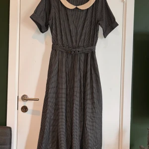 Rutigt långklänning med krage och bälte - Köpt i Japan, märke F.I.N.T.  Size: 36-38.  Snygg långklänning med svartvitt rutigt mönster, korta ärmar och en kontrasterande vit krage i retrostil. Klänningen har ett matchande bälte i midjan som ger en markerad siluett. Perfekt för dig som gillar vintage vibes och klassisk stil.