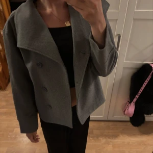 Grå rock/kappa med bred krage - Säljer en stilren grå rock/kappa med bred krage och dubbelknäppning framtill. Jackan har en rak, lite boxig passform och är perfekt för dig som gillar minimalistisk stil. Materialet känns mjukt och ger en clean look. Passar till allt och är lätt att matcha.
