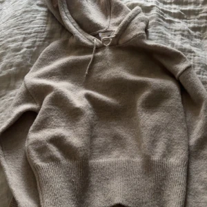 Beige stickad hoodie i ullblandning - Mysig beige hoodie med huva och snörning, stickad i mjuk ullblandning. Tröjan har ribbade muddar vid ärmslut och nederdel. Perfekt för sommarkvällar och inför höst/vinter🥰