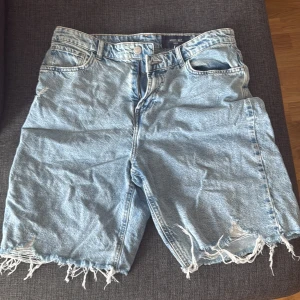 Ljusblå jeansshorts från Noisy May - Säljer ett par ljusblå jeansshorts från Noisy May i storlek Large. De har råa, fransiga benslut och klassisk femficksdesign. Perfekt för dig som gillar en avslappnad och trendig look. Materialet är denim och passformen är normal.