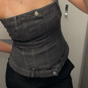 Svart jeanskorsett med knappar - Säljer en svart korsettopp i jeansmaterial med coola knappdetaljer och bältesöglor. Toppen är axelbandslös och har en tight passform som framhäver midjan. Perfekt för dig som vill sticka ut med en edgy vibe.