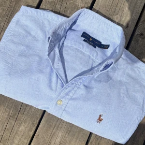 Ralph Lauren Skjorta  (pris diskuterbart) - !! NY SKICK (förutom ett litet märke längst ner som knappt syns) !!Klassisk ljusblå skjorta från Ralph Lauren i SLIMFIT (står custom fit men sitter som slim)  med button-down krage och broderad logga på bröstet. Skjortan har lång ärm, knappar framtill och är tillverkad i mjuk bomull. Perfekt för en clean och stilren look.