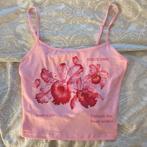 Rosa blommig linnetopp med tryck - Supersöt rosa linnetopp med smala axelband och blommigt tryck framtill i rött och rosa. Texten 'SINCE 1989', 'SIMPLE JOY' och 'Unleash the beast within' ger toppen en unik vibe. Perfekt för dig som gillar färg och statement-plagg.