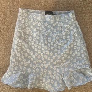 Blommig ljusblå kjol från Pieces - Supersöt ljusblå kortkjol från Pieces med vita blommor och volang nertill. Kjolen har en feminin och fräsch look, perfekt för dig som gillar romantiska vibes. Materialet känns lätt och somrigt, och mönstret ger en extra touch till outfiten.