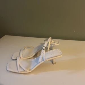 Vita skor med klack från H&M - Snygga vita sandaler från H&M med smala remmar och fyrkantig tå. Skorna har en låg klack och justerbart spänne vid ankeln. Perfekta för sommarens alla tillfällen och ger en clean och trendig look.