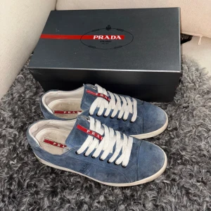 Blå sneakers från Prada - Box osv följer med. Snygga blå sneakers från Prada i mocka med vita snören och en diskret logga på sidan. Perfekta för en stilren look med en touch av lyx.                                         ! Skorna är använda och har en del slitage men är i fullt acceptabelt skick !