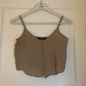 Beige linnetopp med knappar från Bik Bok - Snygg beige linnetopp från Bik Bok med smala axelband och v-ringning. Toppen har dekorativa tygklädda knappar framtill och är croppad i modellen. Perfekt att styla med jeans eller kjol för en trendig look.