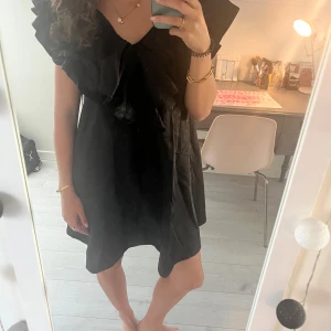 Svart kortklänning med volang - Supersnygg svart kortklänning med v-ringning och stora volangdetaljer över axlarna. Klänningen har en lös passform och är tillverkad i ett glansigt material.