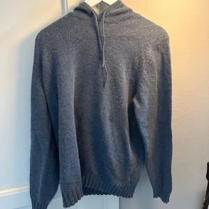 Blå stickad hoodie med huva - Mysig ljusblå stickad hoodie med huva och dragsnöre. Tröjan har långärmad modell och ribbade muddar vid ärmslut och nederkant. Perfekt för kyliga dagar när du vill vara både bekväm och snygg.