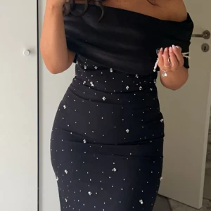 Svart långklänning  - Säljer en svart långklänning med offshoulder-modell och tight passform. Klänningen är dekorerad med små pärlor över hela tyget och har ett stort pärlbroderat  blomdetalj nedtill. Perfekt för dig som vill sticka ut med elegans. Haft den 1 gång och beställd från Tyskland - gott skick. Ny pris 3800