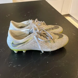 Nike Hypervenom fotbollsskor grå/gula - Nike Hypervenom fotbollsskor i ljusgrått med stora silvriga Swoosh-loggor och gula detaljer. Skorna har snörning och platt sula med fasta dobbar för spel på gräs. Ovandelen är i syntetmaterial med textur för bättre bollkontroll. Perfekta för dig som vill sticka ut på planen.