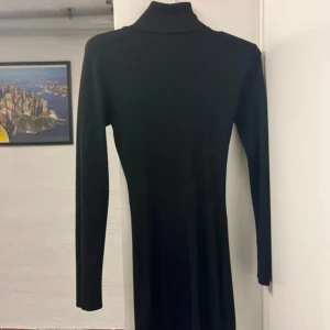 Svart ribbad långklänning med polokrage - En svart, ribbad långklänning med polokrage och lång ärm. Klänningen har en slim passform och är tillverkad i ett stickat material som ger en snygg siluett. Perfekt för dig som gillar en stilren och enkel look.