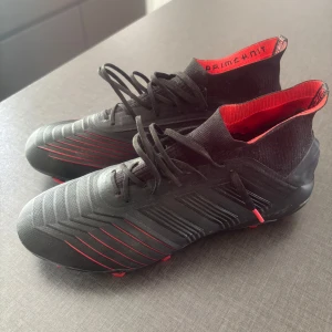 Adidas Predator fotbollsskor svart/röd - Säljer ett par Adidas Predator fotbollsskor i svart med röda detaljer och mönstrad ovandel. Skorna har snörning, hög strumpliknande krage i Primeknit-material och dobbar för bra grepp på planen. Perfekta för dig som vill sticka ut på matchen!