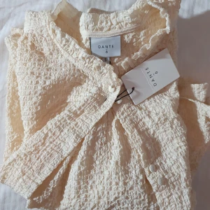 Ljus kortklänning från Dante6 - Supersöt ljus kortklänning från Dante6 med struktur i tyget och fina knappar framtill. Klänningen har lång ärm och lätt volang vid halsen. Perfekt för dig som gillar en stilren och chic look med en twist.