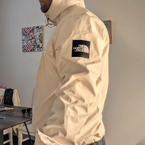 Beige vindjacka från The North Face - Säljer en beige vindjacka från The North Face med huva och logga på ärmen. Jackan har dragsko vid huvan och är perfekt för blåsiga dagar. Snygg och enkel design som funkar till det mesta. Passar dig som gillar streetwear och outdoor vibes.