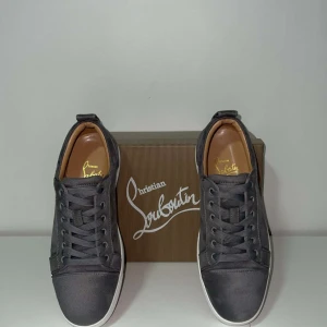 Gråa sneakers från Christian Louboutin - Snygga gråa sneakers från Christian Louboutin i mocka med klassisk vit sula och ikonisk röd undersida. Skorna har rund tå, snörning och diskreta detaljer. Perfekta för dig som vill ha en lyxig och stilren look med streetkänsla.