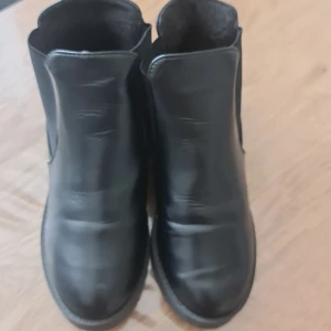 Svarta chelsea boots i skinn - Stilrena svarta chelsea boots med grov sula och låg klack. Skorna har elastiska paneler på sidorna för enkel påtagning och är tillverkade i slätt skinn. Perfekta för dig som vill ha en klassisk och edgy look till höst och vinter.