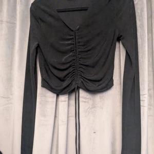 Svart långärmad topp Noisy May - Svart långärmad topp från Noisy May med snygg v-ringning med snörning framtill som ger en rynkad effekt. Modellen är croppad, Lite glittrig i materialet och använd fåtal gånger 🫶🏻