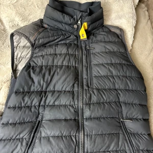 Svart dunväst från Parajumpers - Snygg svart dunväst från Parajumpers med quiltad design och hög krage. Västen har flera dragkedjeförsedda fickor, en gul detalj vid halsen och patch med logga på ryggen. Insidan har märkets klassiska etikett och orange 'Security Pocket'-detalj.