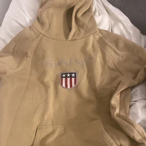 Beige hoodie från GANT med brodyr - Snygg beige hoodie från GANT med broderad logga och sköld på bröstet. Tröjan har huva, känguruficka och är tillverkad i mjuk bomull. Perfekt för dig som gillar klassisk och stilren streetwear.