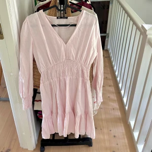 Ljusrosa kort klänning från Zara - Supersöt ljusrosa klänning från Zara med v-ringning, smock i midjan och långa ärmar med resår vid handlederna. Klänningen har volangdetaljer nertill och är tillverkad i ett mjukt bomullstyg. Perfekt för vår och sommar!