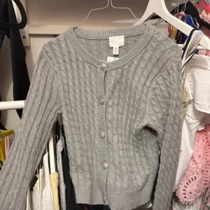 Grå kabelstickad kofta från H&M - Supersöt grå kabelstickad kofta från H&M i storlek XS. Koftan har rund halsringning, knappar framtill och ribbade muddar. Perfekt att slänga över en topp för en chill look. Materialet är mjukt och skönt, och den är långärmad för extra mysfaktor.