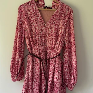 Rosa blommig klänning med bälte - Supersöt rosa blommig klänning med v-ringning, långa puffärmar och ett smalt svart bälte i midjan. Klänningen har ett flowy fall och små knappar framtill. Perfekt för dig som gillar romantiska vibes och vill sticka ut lite extra.