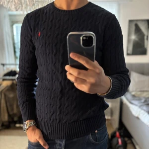 Mörkblå kabelstickad tröja Polo Ralph Lauren - Säljer en mörkblå kabelstickad tröja från Polo Ralph Lauren i storlek XL (Barnstorlek) = S/M. Tröjan har rund halsringning, långa ärmar och den klassiska röda broderade loggan på bröstet. Perfekt för dig som gillar en stilren och tidlös look.
