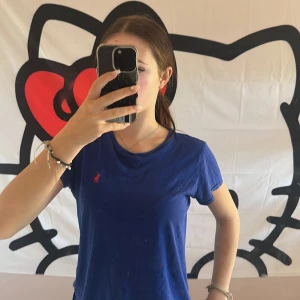 Blå t-shirt från Ralph Lauren - Blå t-shirt från Ralph Lauren med klassisk passform och korta ärmar. T-shirten har rund halsringning och den ikoniska rosa Polo-loggan broderad på bröstet. Tillverkad i mjuk bomull som är skön mot huden. Perfekt till jeans eller shorts. Aldrig använd!