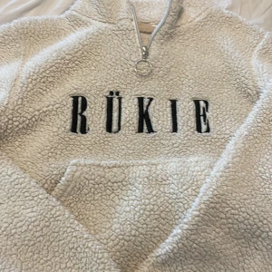 Vit teddytröja med half zip från RÜKIE - Supermjuk och fluffig vit teddytröja från RÜKIE med svart broderad logga framtill. Tröjan har en snygg half zip med ringdetalj och stor känguruficka. Perfekt för kyliga dagar när du vill vara både varm och trendig.
