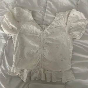 Vit blus med puffärm och volang - Superfin vit blus med broderade blommor, puffärmar och v-ringning. Toppen har smockad detalj framtill och volang nertill som ger en söt och trendig look. Perfekt till jeansshorts eller kjol för en fräsch sommarstil.