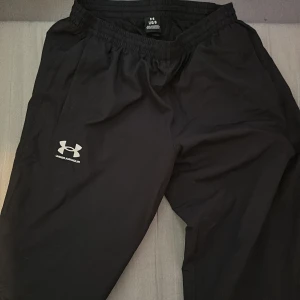 Svarta träningsbyxor Under Armour L - Svarta träningsbyxor från Under Armour med elastisk midja och logga på benet. Lätta och bekväma, perfekta för gymmet eller löprundan. Tillverkade i syntetmaterial som andas och torkar snabbt. Skador på högra sidan vid låren. Ingen Frakt